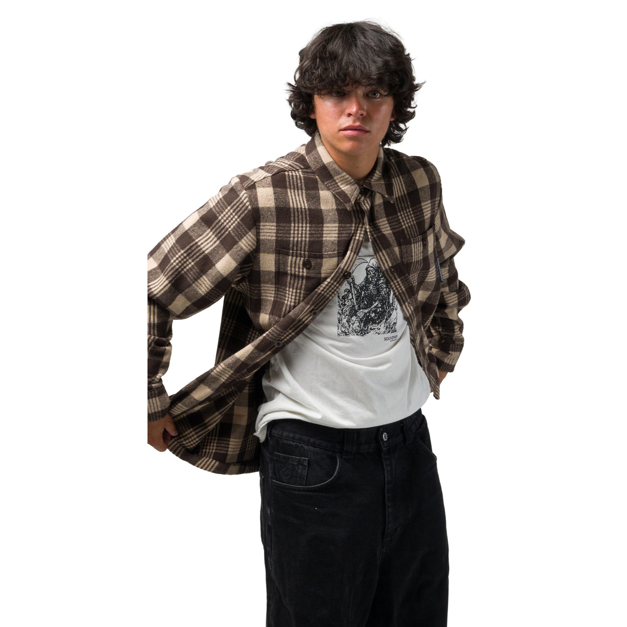 A model wearing a brown and beige plaid souvenir flannel shirt layered over a white graphic t-shirt and black jeans, posed against a solid white background.