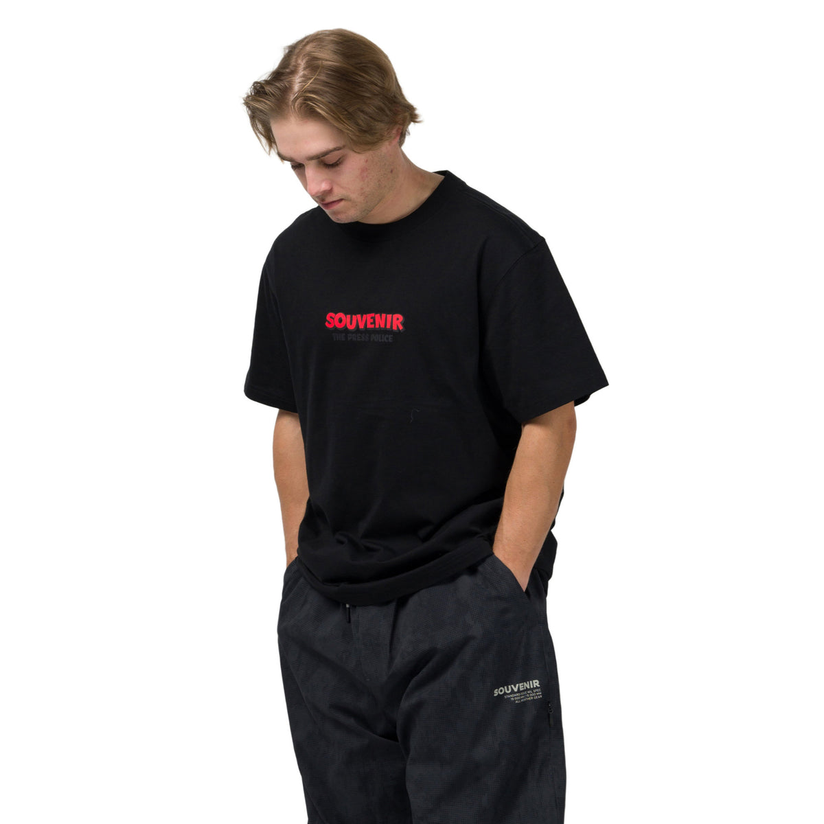A man wearing a black t-shirt with red 'SOUVENIR' text on the chest and black pants, standing against a plain white background.