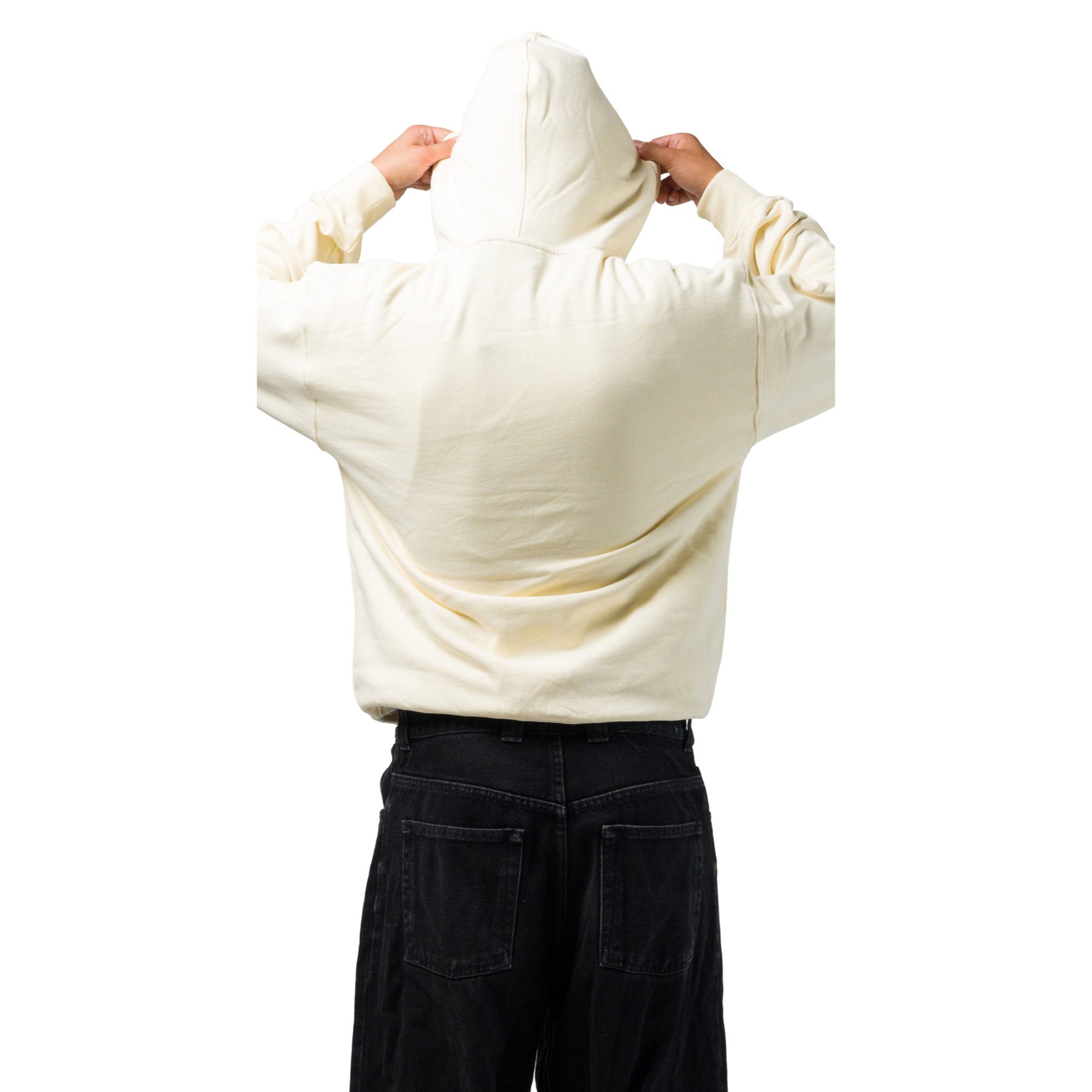 A person viewed from behind wearing a cream-colored oversized hoodie and dark jeans, with both hands adjusting the hood against a plain white background.
