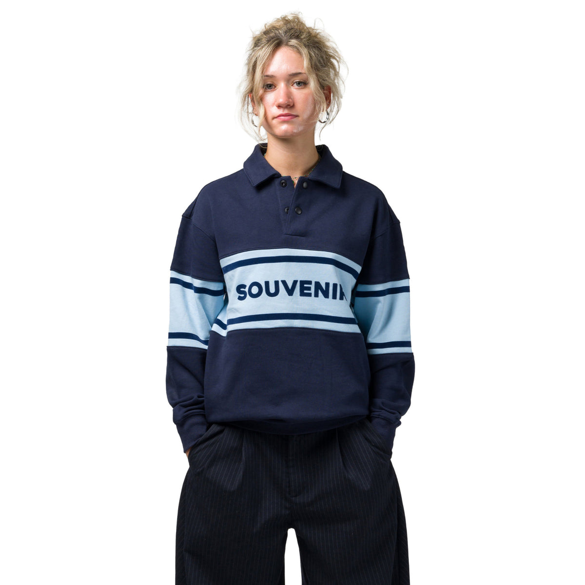 A model wearing a navy blue long-sleeve rugby style Souvenir shirt with light blue and white stripes across the chest and sleeves, featuring the word 'SOUVENIR' printed in navy on the chest, paired with dark pinstriped trousers against a solid white background.