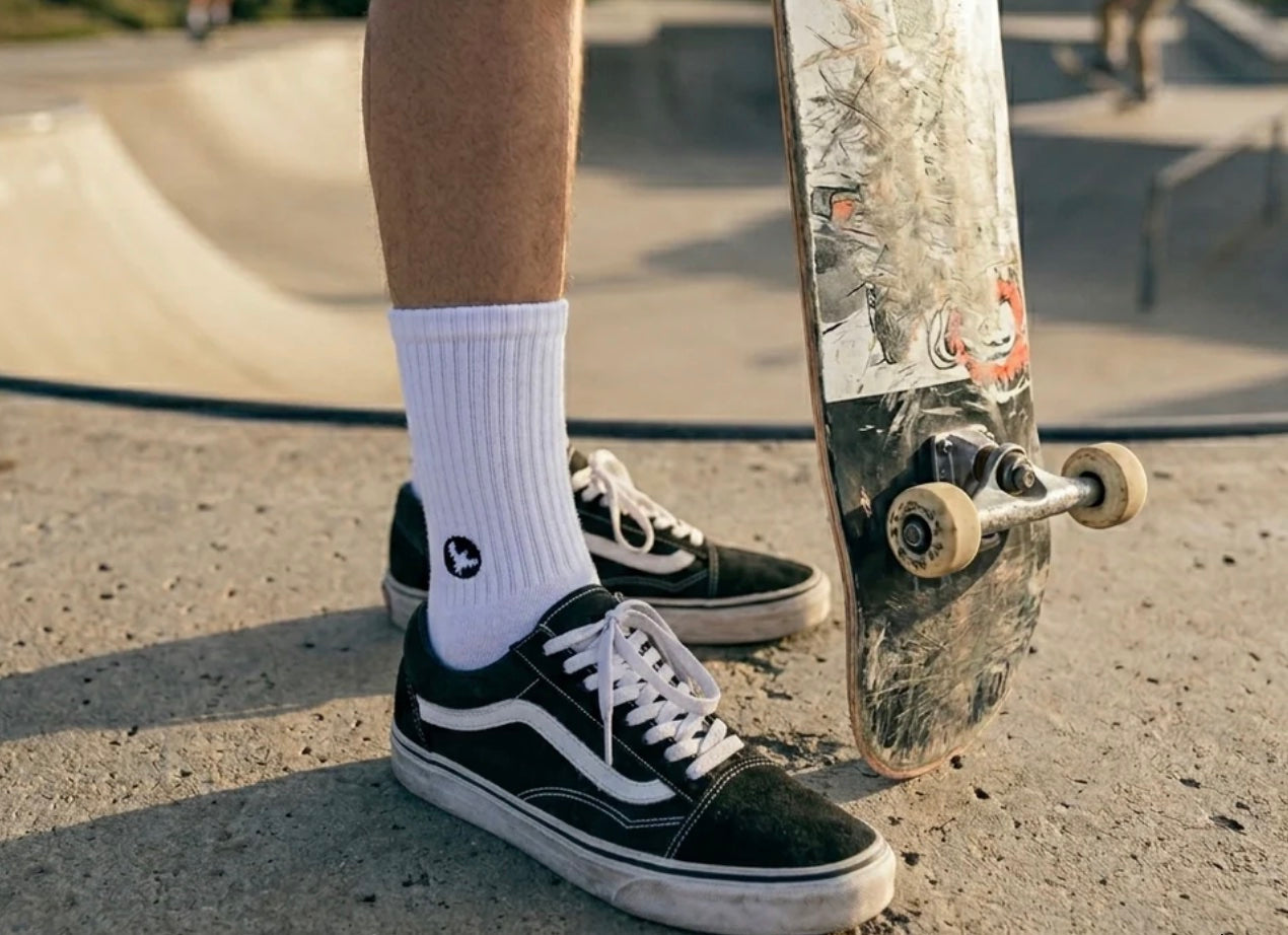 Person wearing black Vans sneakers with white socks, holding a skateboard at a skate park.
