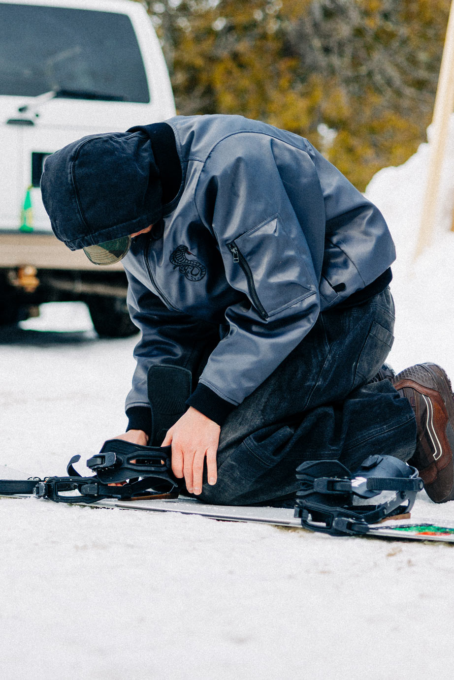 Rider wearing the Souvenir 3 Ply Denim Pant 2027 in Black Denim while kneeling in the snow and adjusting snowboard bindings.