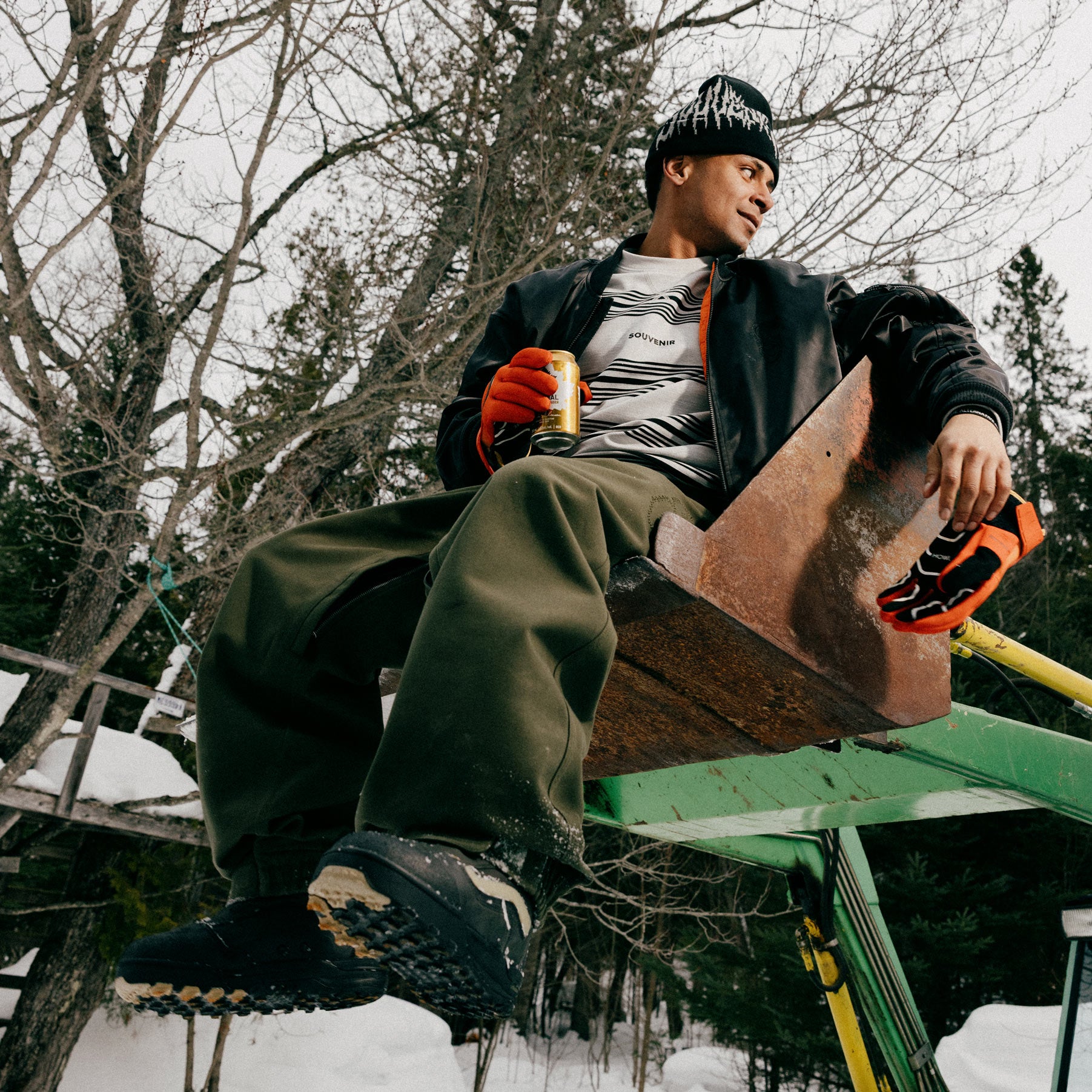 Rider wearing the Souvenir 3 Ply Workwear Pant 2027 in Dark Olive Oxford while seated outdoors in the snow, showing the relaxed wide-leg fit.