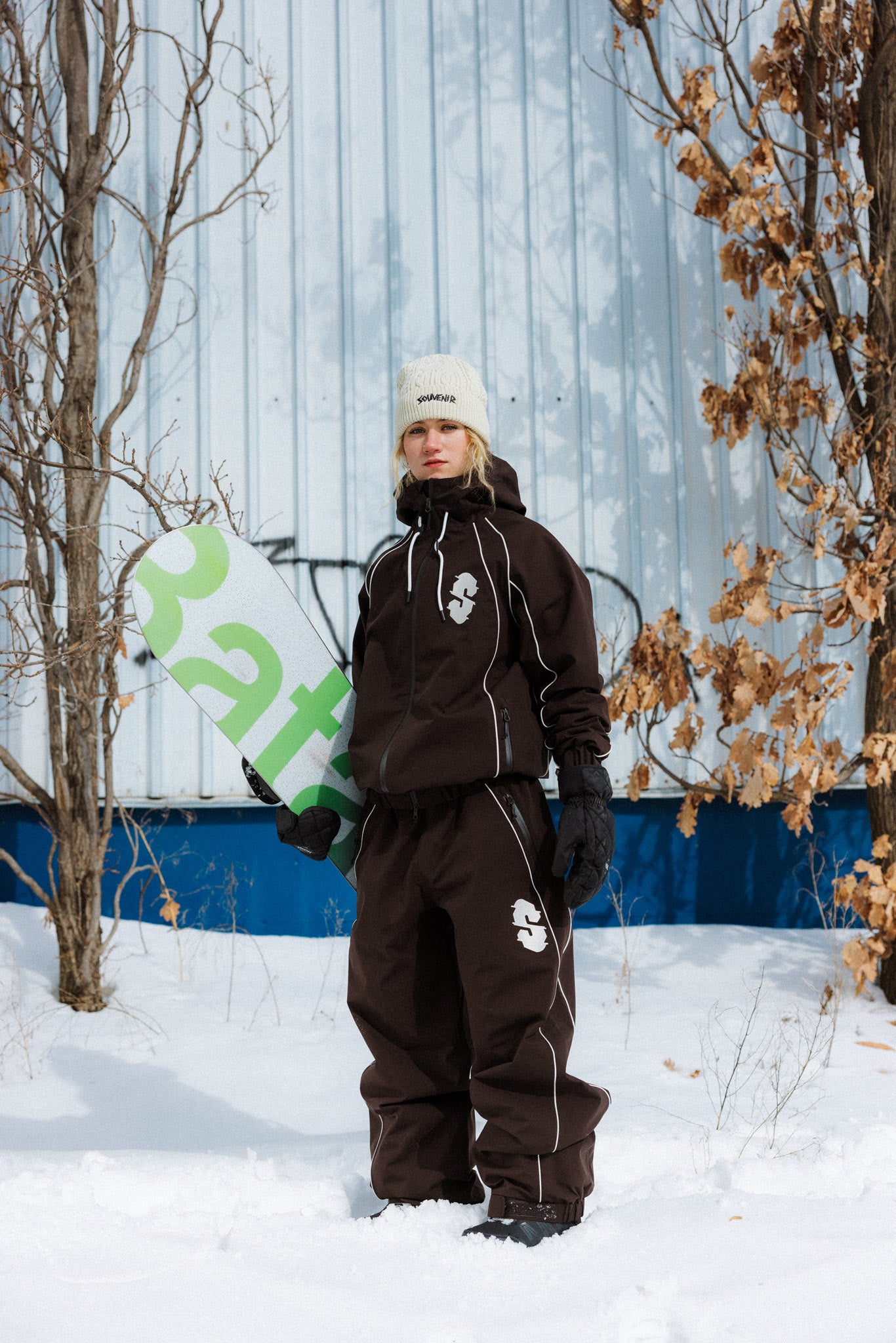 Snowboarder wearing the Souvenir Insulated Track Pant and Jacket 2027 in Cocoa while holding a snowboard in the snow, showing the full baggy track silhouette.