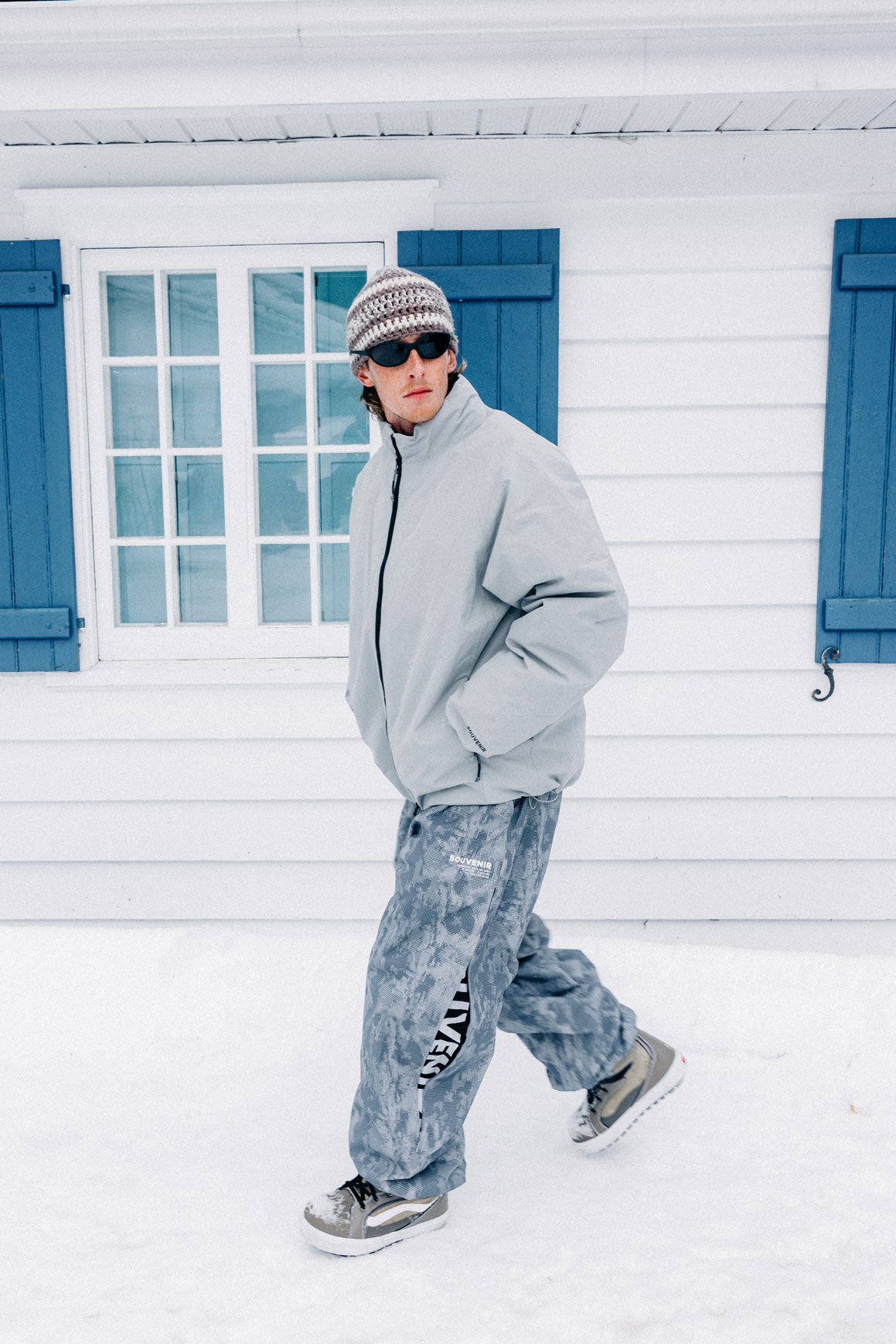 Snowboarder wearing the Souvenir Parachute Pant 2027 in Cool Grey Blotch Print with a grey jacket and snowboard boots in the snow.