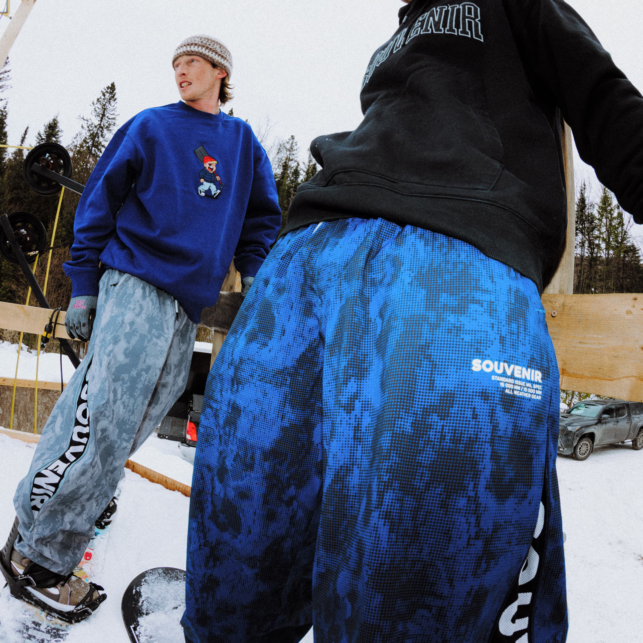 Snowboarders wearing Souvenir Parachute Pants in Petrol Blue and Cool Grey Blotch Print standing together in the snow.