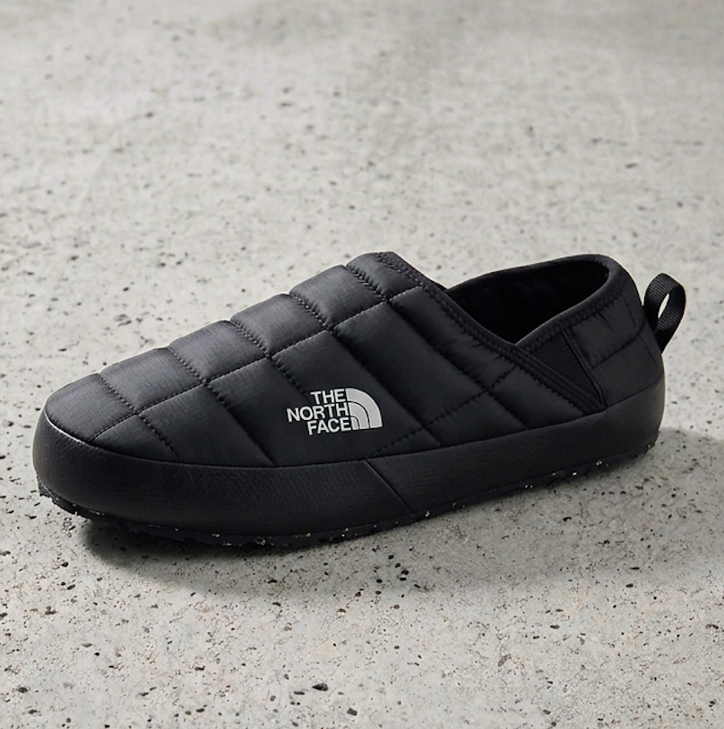 A black quilted slip on shoe by The North Face, shown from a side angle on a textured gray concrete surface.