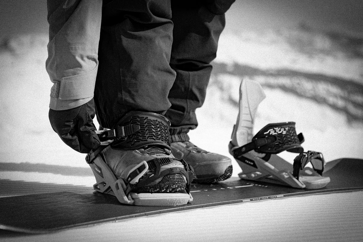 Black and white close-up of a snowboarder strapping a boot into snowboard bindings on a snowboard, used for the Milosport Men’s Snowboard Bindings collection.