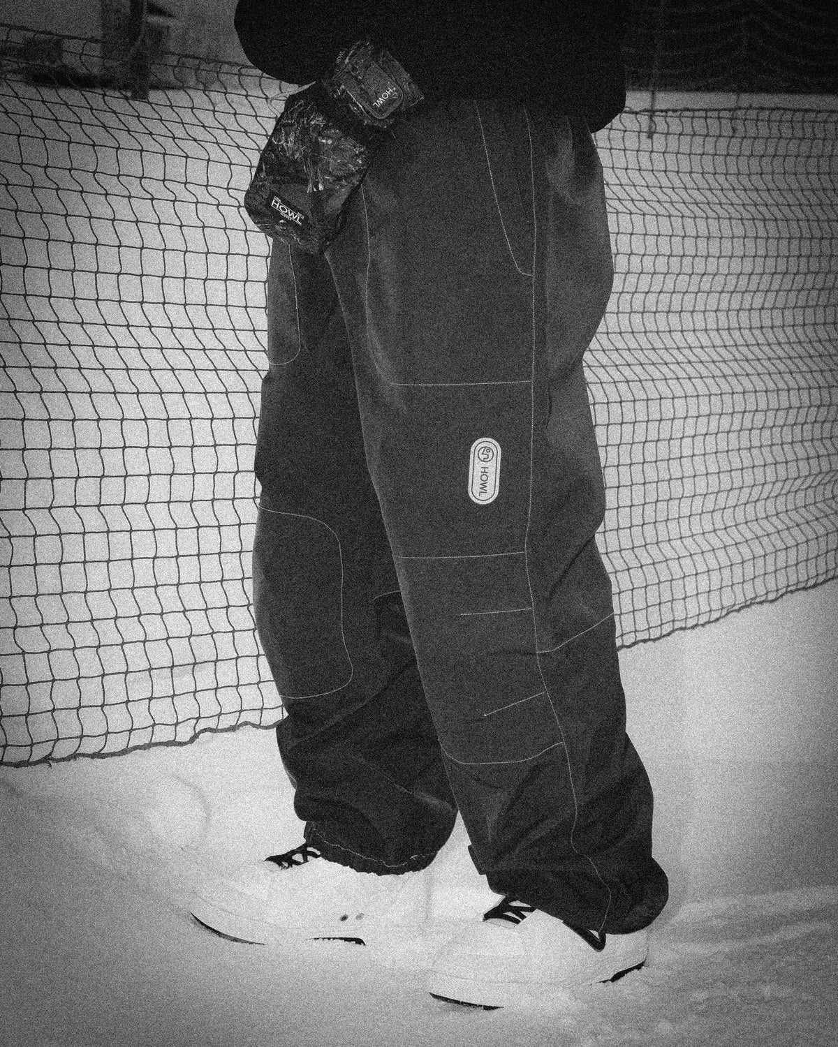 Black and white lifestyle image of a snowboarder wearing brown snow pants and white snowboard boots on snow, used for the Milosport Snow Pants collection.