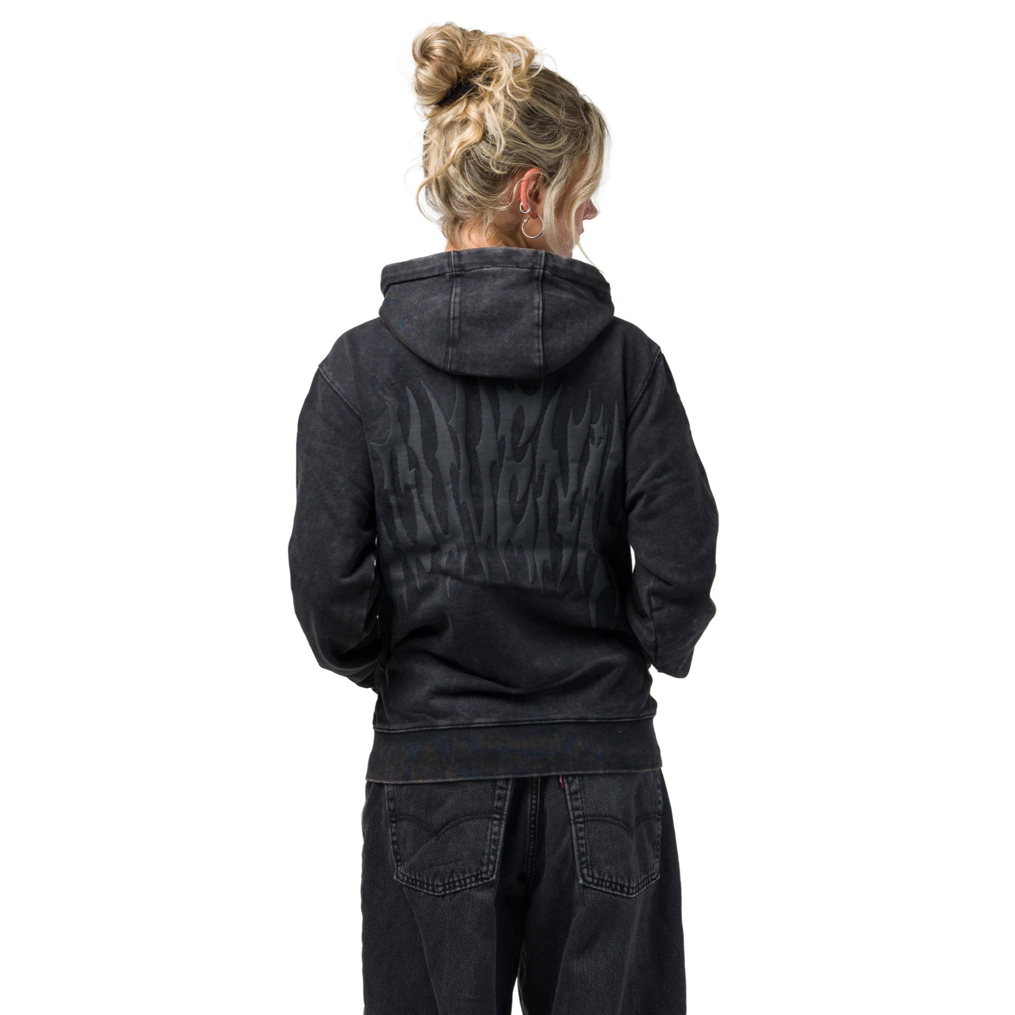 A woman viewed from behind wearing a faded black washed souvenir hoodie with a tonal flame graphic on the back, paired with dark jeans, against a solid white background.