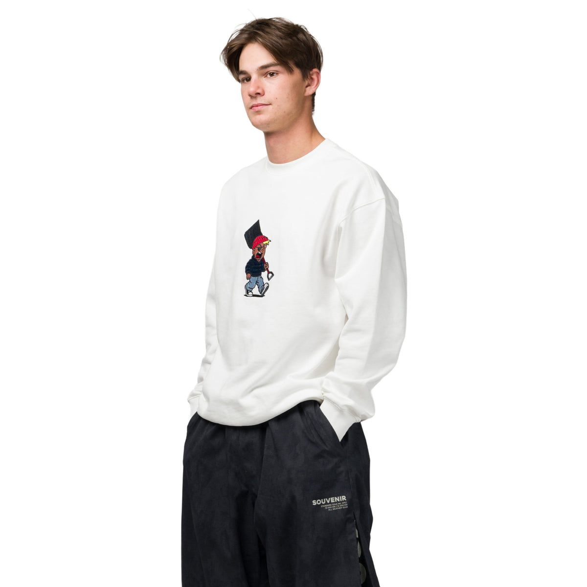 A young man wearing a white crewneck sweatshirt with a small cartoon graphic on the chest, paired with black cargo pants, set against a plain white background.