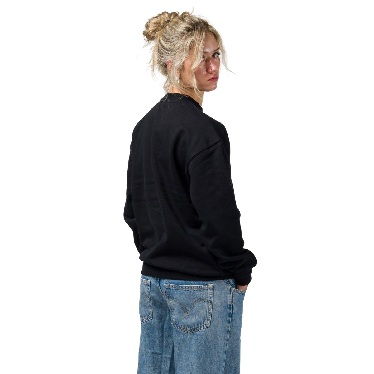 A woman with blonde hair in a bun is shown from a side back angle wearing a black crewneck souvenir sweatshirt and blue jeans against a solid white background.