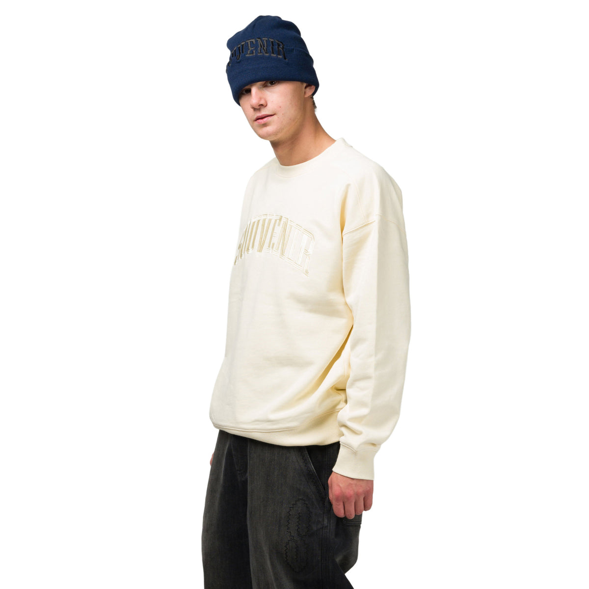 A model wearing a cream-colored crewneck sweatshirt with a tonal embroidered logo, paired with dark jeans and a navy blue beanie, set against a plain white background.