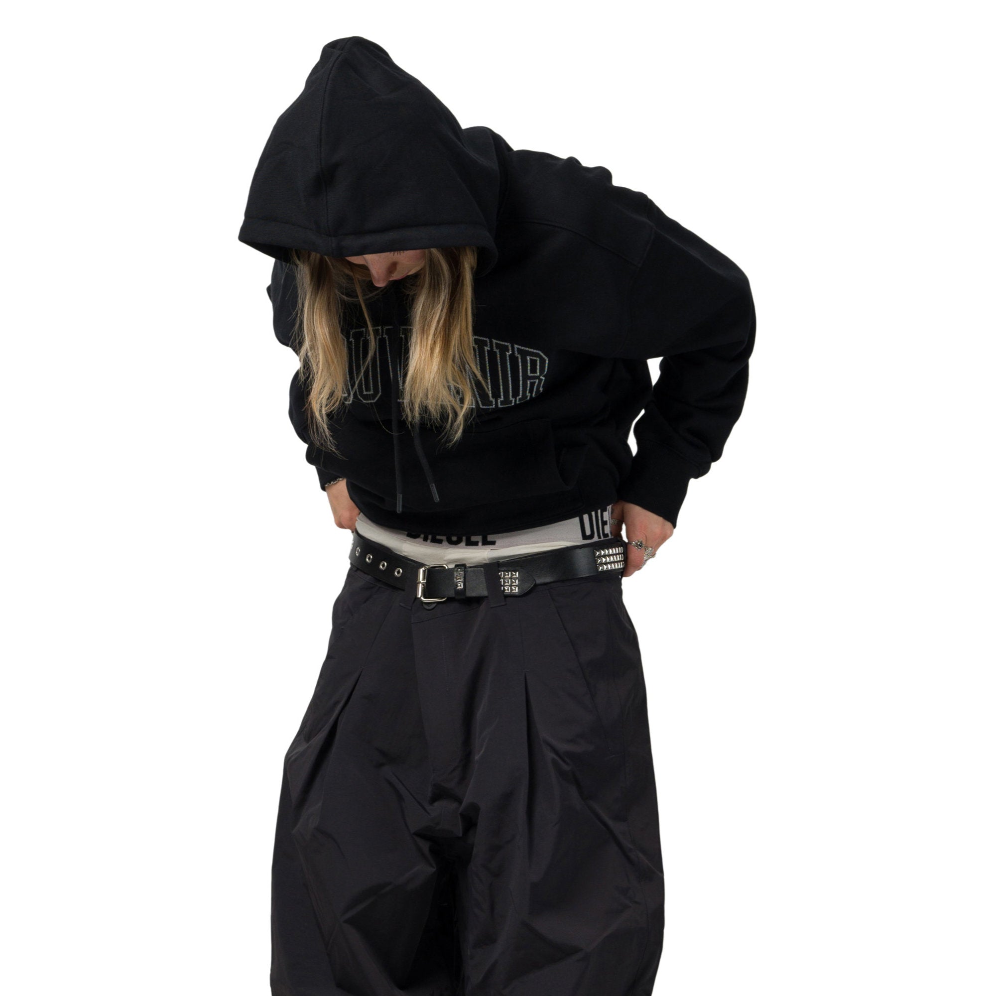 A person wearing a black oversized hoodie with a graphic print, black pleated pants, and a black studded belt, shown against a plain white background.