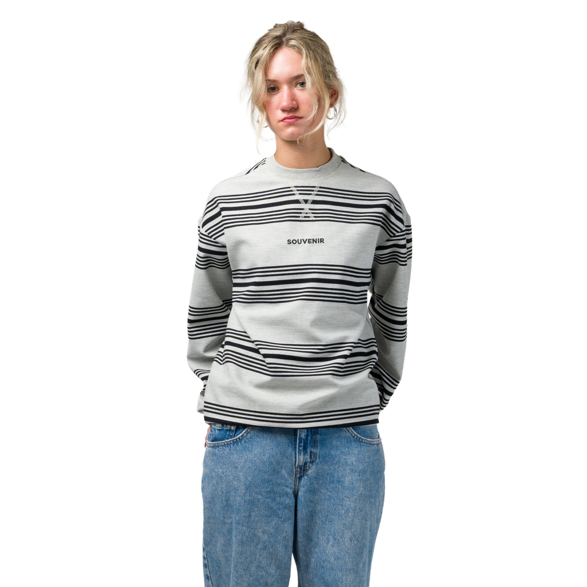 A woman modeling a light gray crewneck souvenir sweatshirt with black horizontal stripes and 'SOUVENIR' printed on the chest, paired with blue jeans against a plain white background.