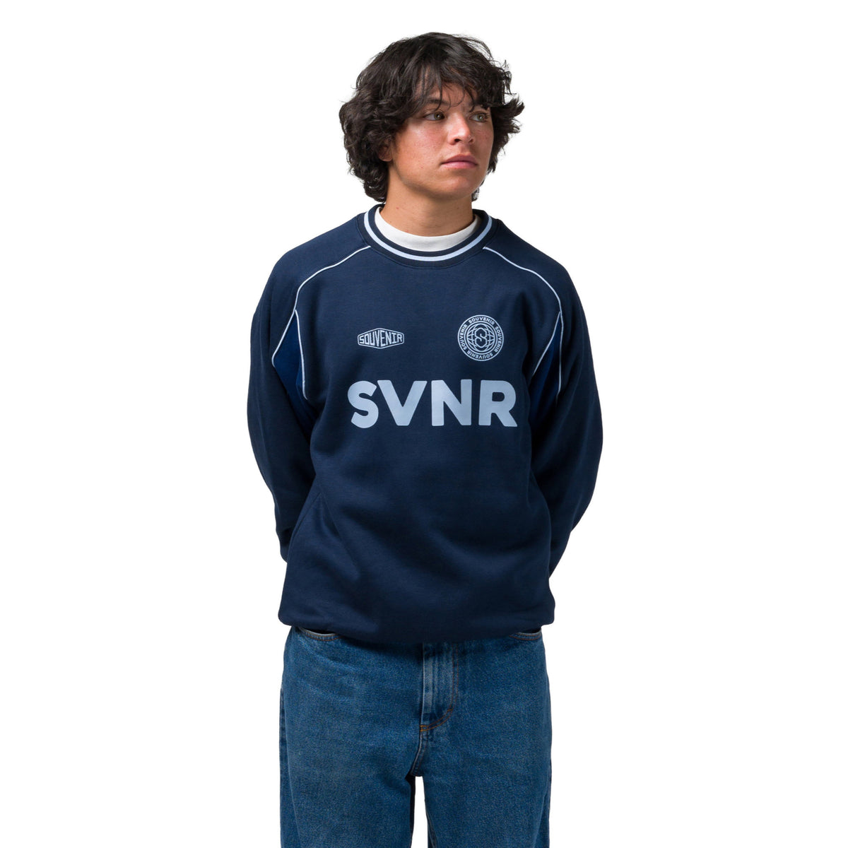 A young man wearing a navy blue sweatshirt with 'SVNR' printed in white across the chest, paired with blue jeans, photographed against a solid white background.