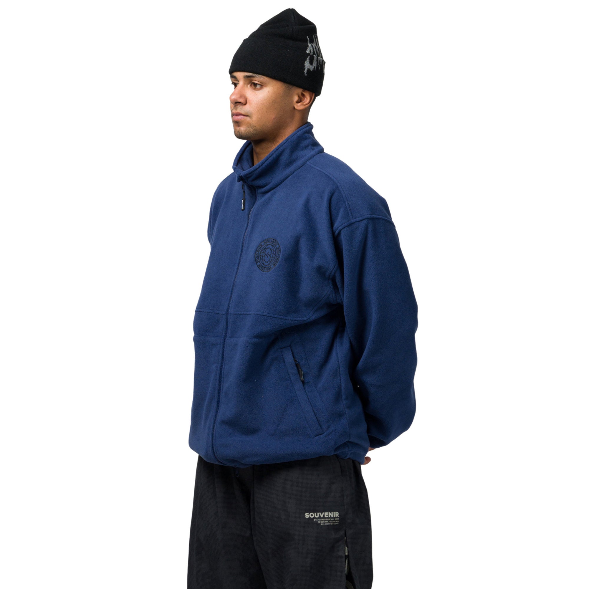A man wearing a navy blue fleece jacket with a tonal circular logo on the left chest, paired with black pants and a black beanie, shown in profile against a white background.