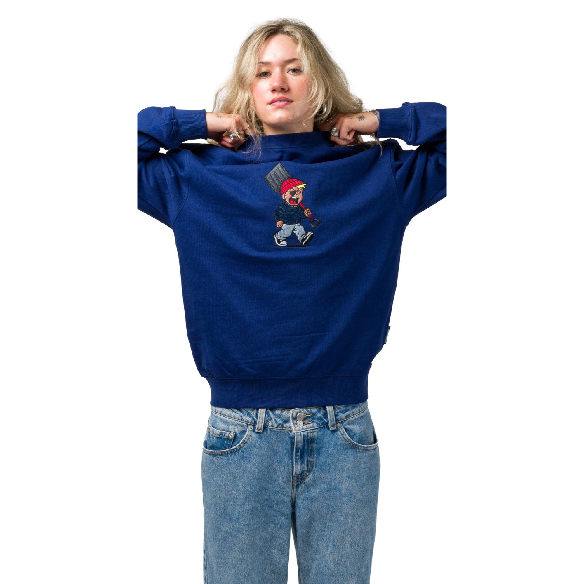 A woman wearing a Souvenir petrol blue crewneck sweatshirt with a small cartoon graphic of a character holding a pickaxe on the chest, paired with light wash blue jeans, against a plain white background.