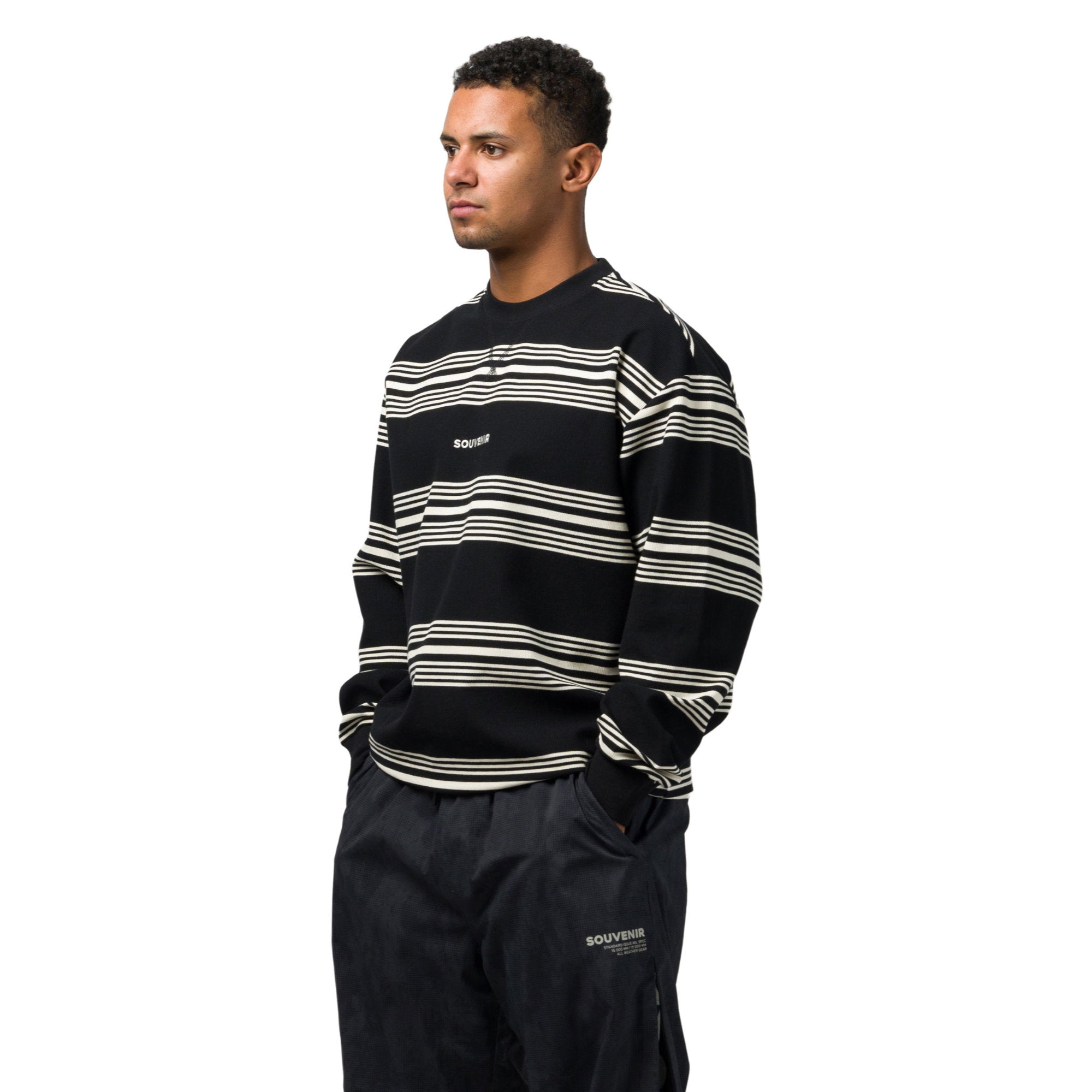 A man wearing a black and off white horizontally striped long sleeve sweatshirt with a small white 'souvenir' logo on the chest, paired with dark cargo pants, photographed against a solid white background.