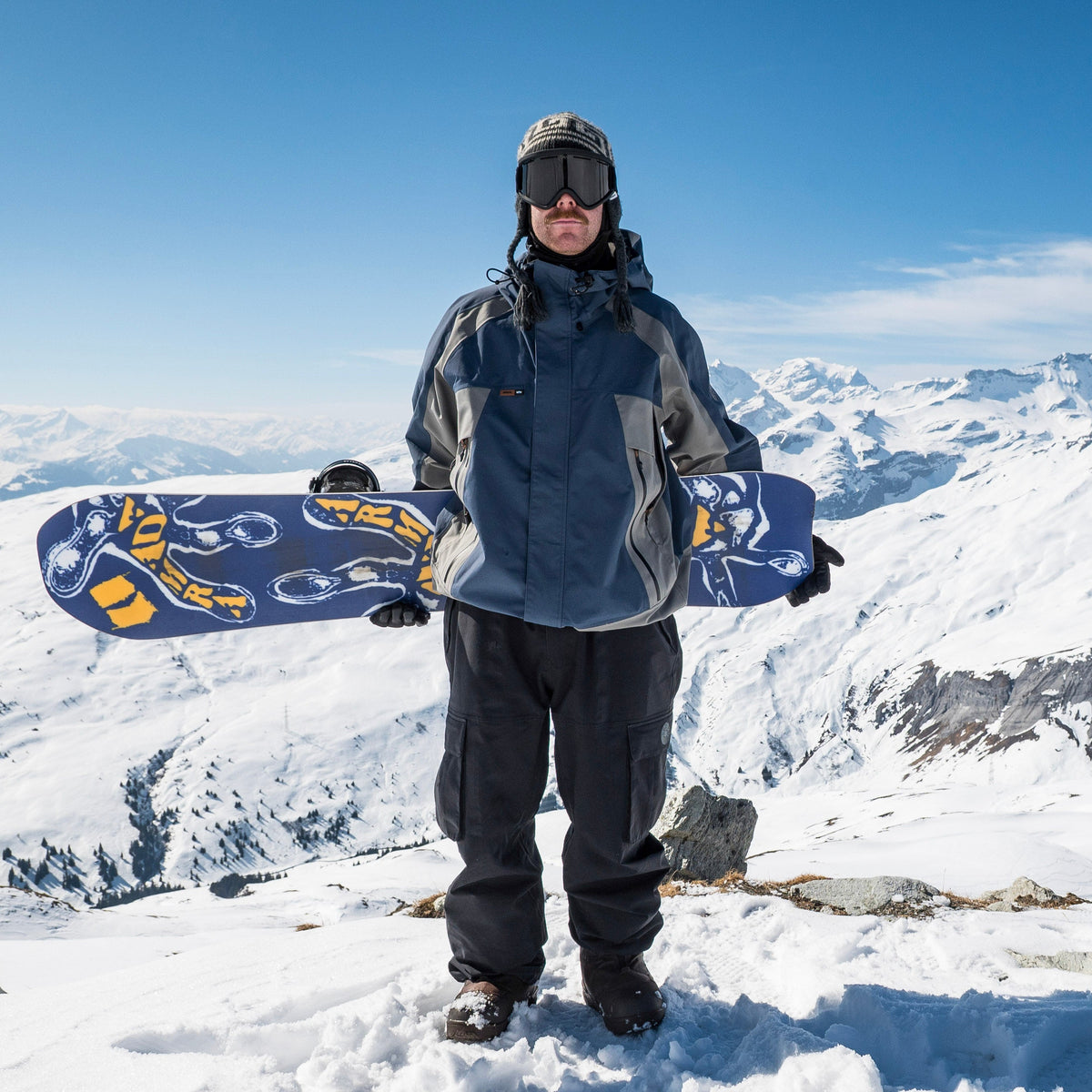 Armada Sicko Snowboard 2027 with Alek standing on snowy terrain, showcasing the board's design and construction.