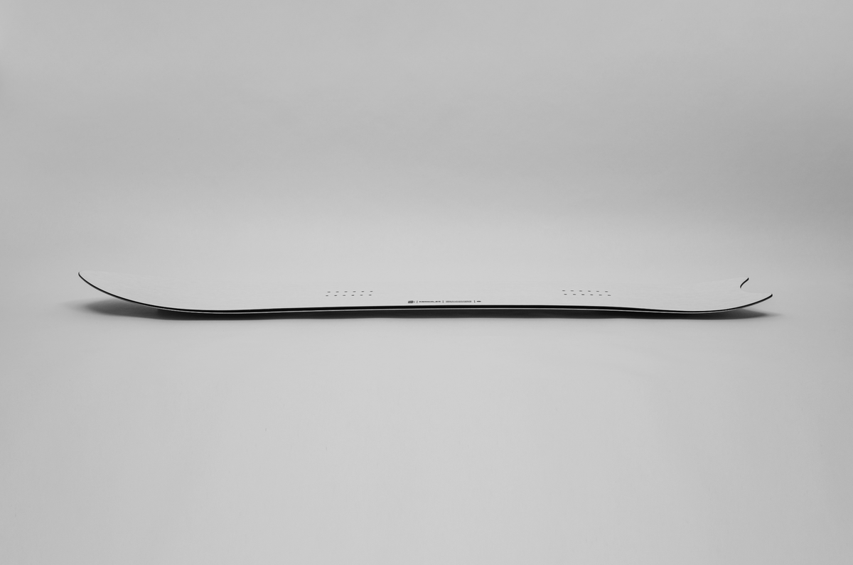 Korua Pencil snowboard in snow, side view showing sleek shape and design, ideal for carving and freestyle snowboarding.