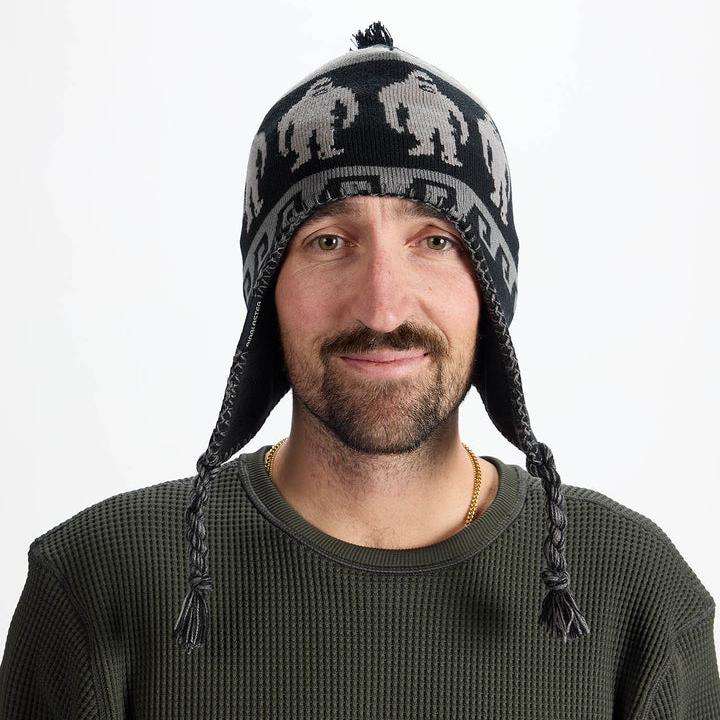 Airblaster Squatchamama Beanie 2027 in Charcoal shown from the front with pom top, ear flaps, braided tassels and Sasquatch knit graphics.