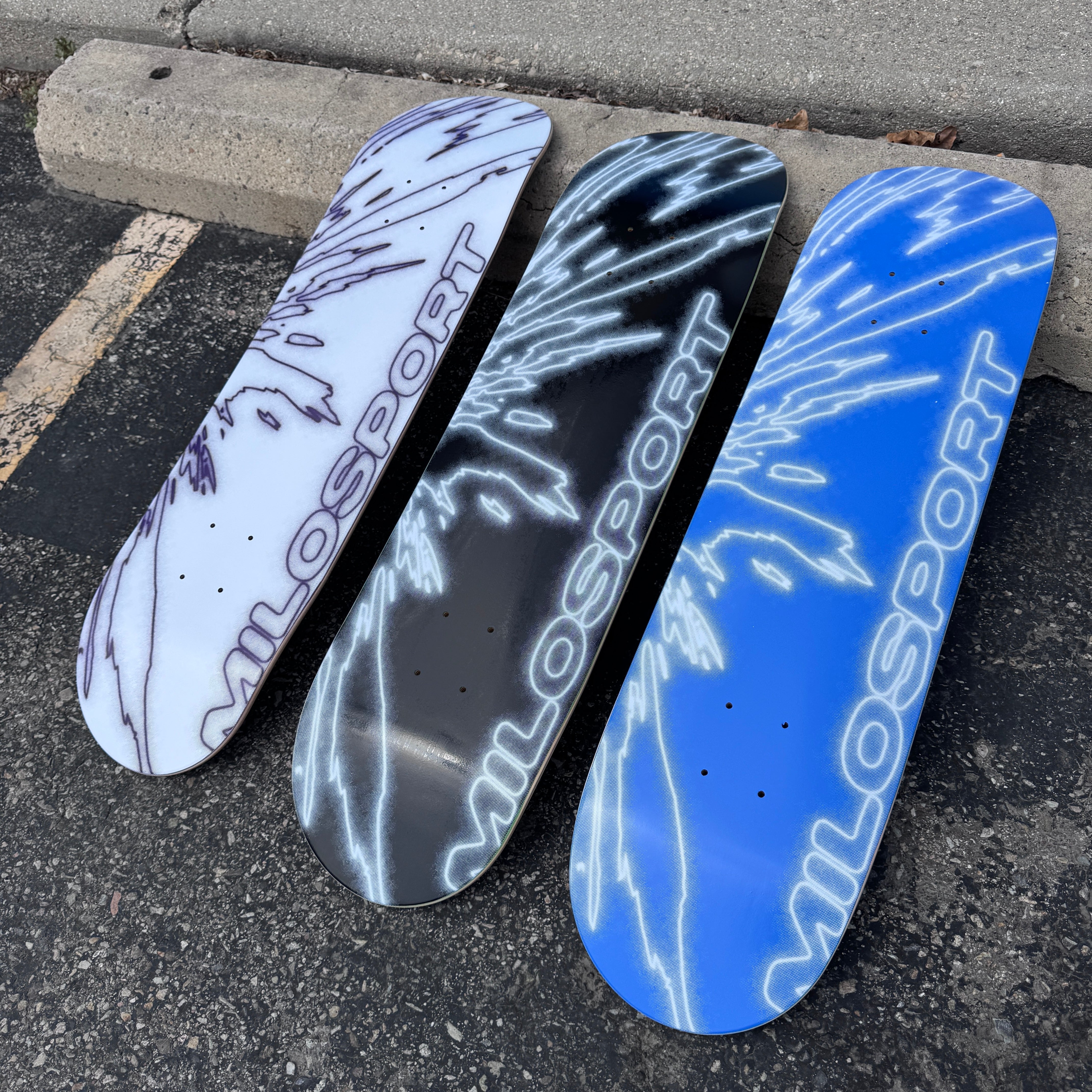 hree Milosport Fracture skateboard decks in white and purple, black and white, and blue and white displayed outdoors on pavement.