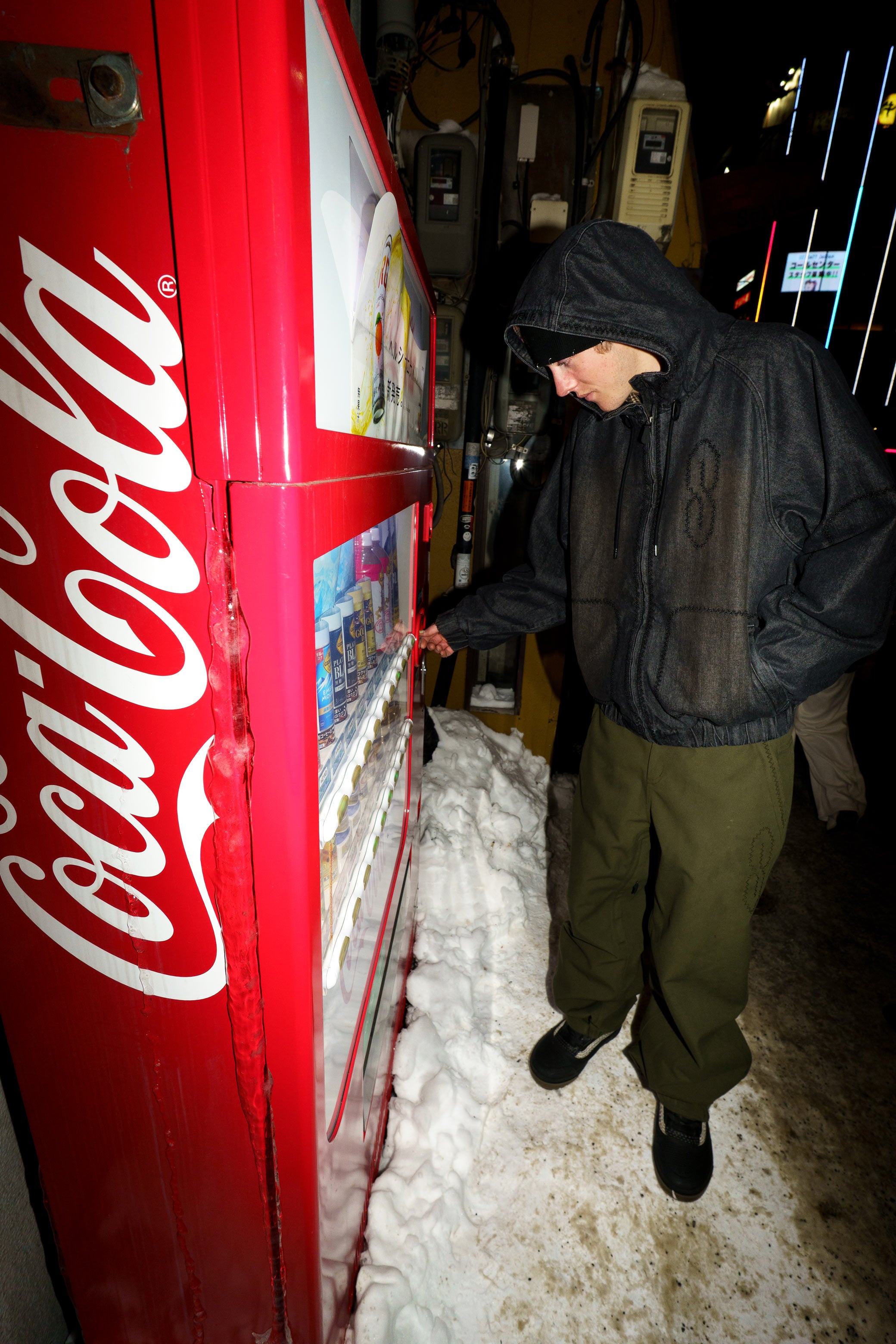 Rider wearing the Souvenir Denim Bomber Jacket 2027 in Black beside a vending machine at night, showing the oversized bomber fit and faded denim finish.