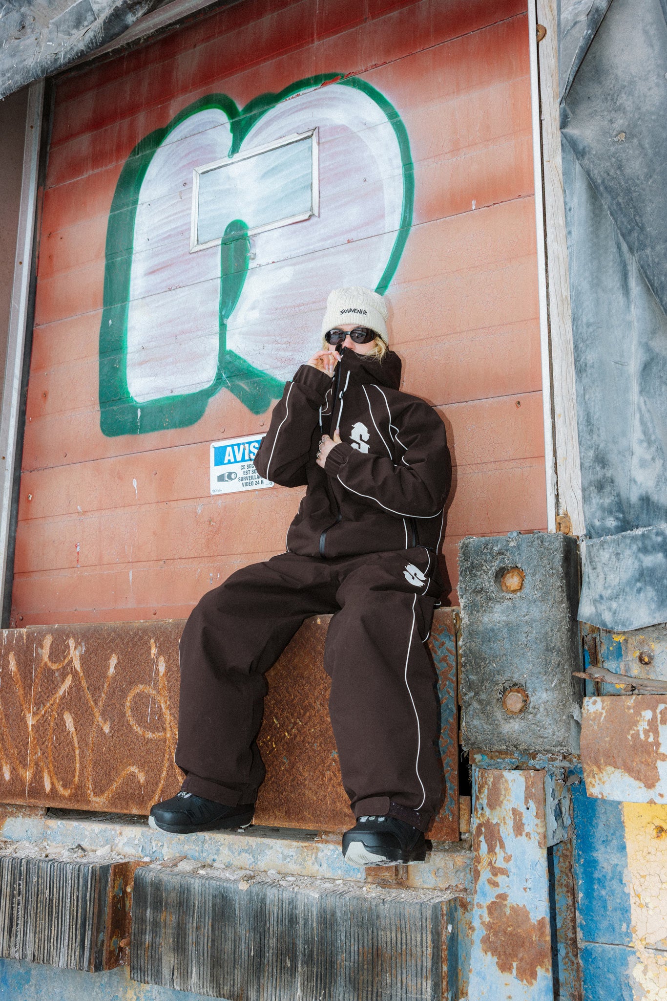 Rider wearing the Souvenir Insulated Track Pant and Jacket 2027 in Cocoa while seated outdoors, highlighting the oversized track fit, reflective piping and Souvenir thigh logo.