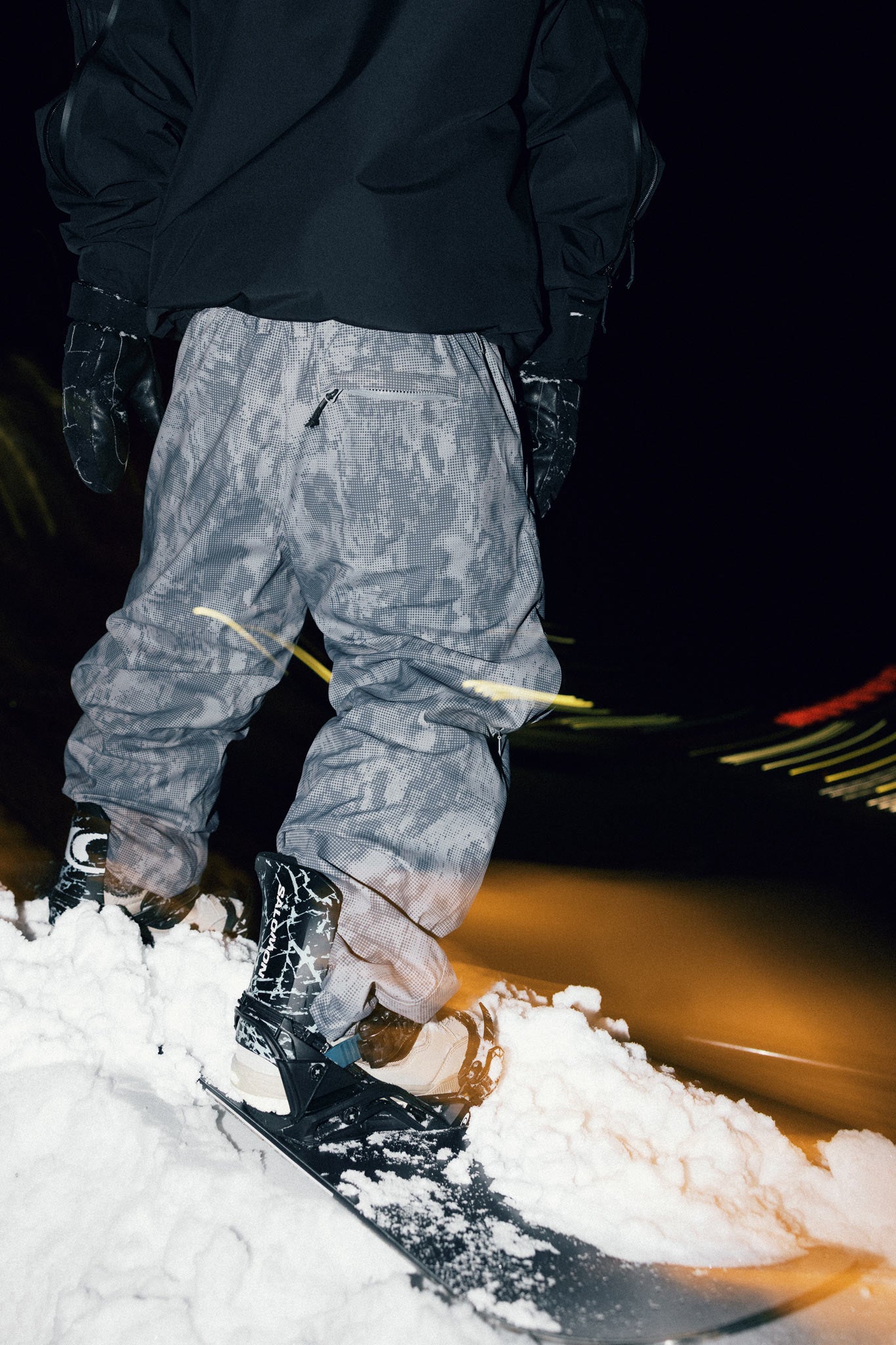 Rear lifestyle view of the Souvenir Parachute Pant 2027 in Cool Grey Blotch Print worn with snowboard boots and bindings.