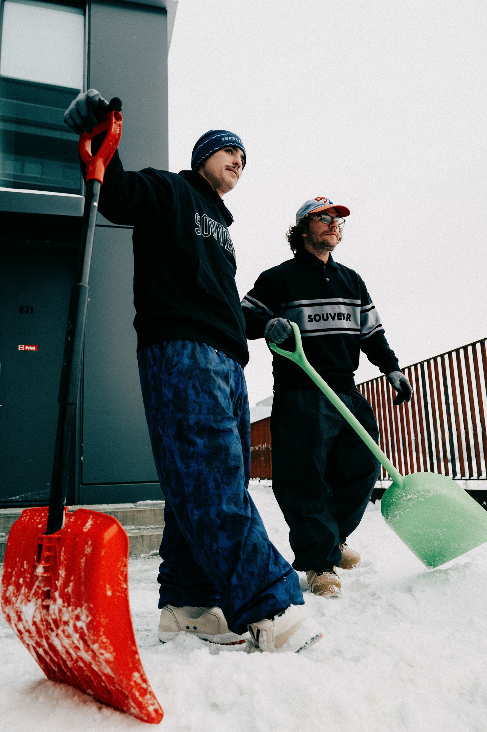 Snowboarder wearing the Souvenir Parachute Pant 2027 in Petrol Blue Blotch Print while carrying a snow shovel.