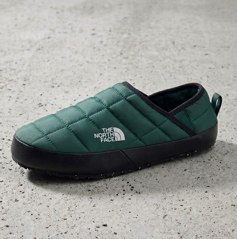 A single quilted slip on shoe by The North Face in dark green with a black sole, shown on a textured gray concrete surface.