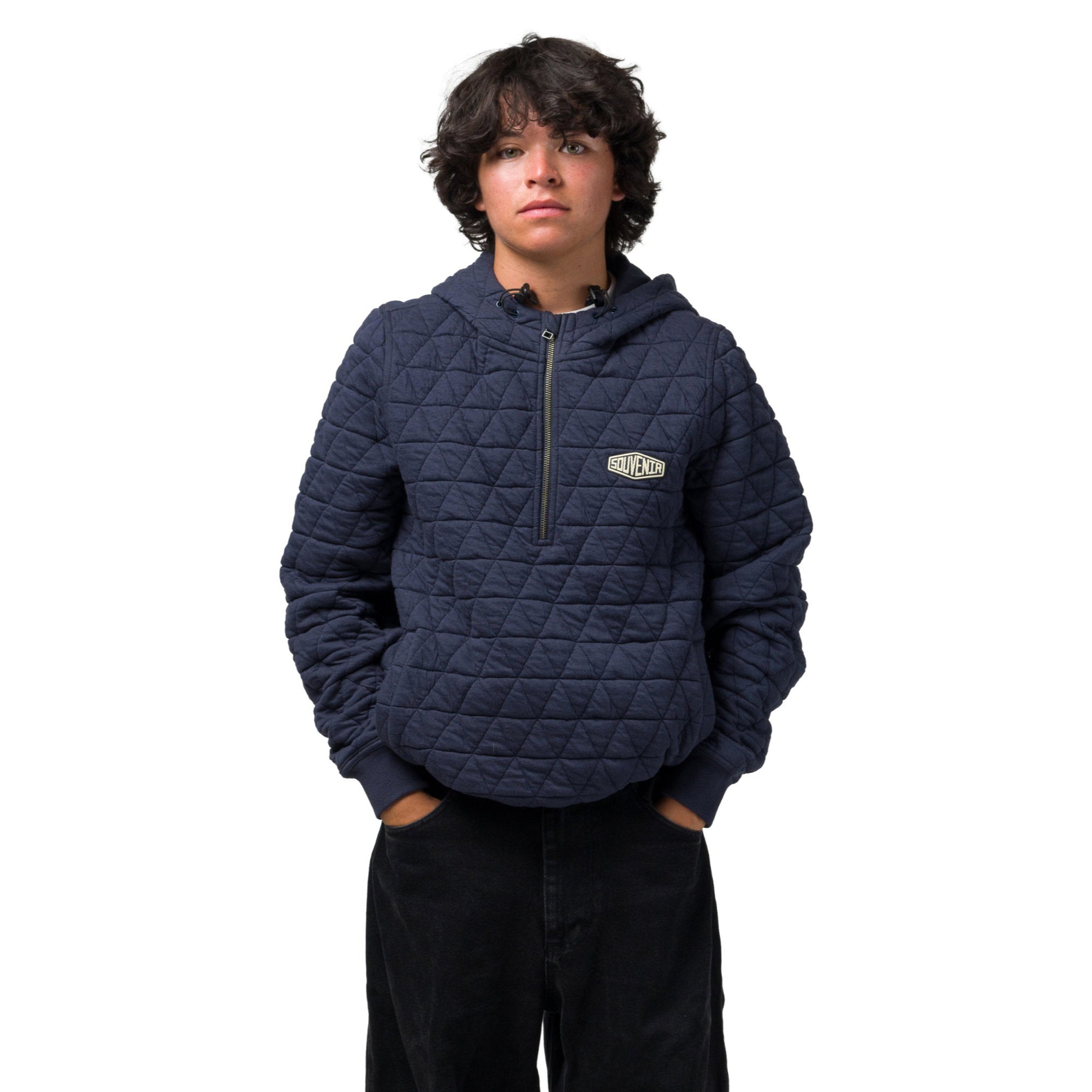 A person wearing a navy blue quilted half-zip hoodie with a small white and black 'souvenir' logo patch on the left chest, paired with dark pants, against a plain white background.