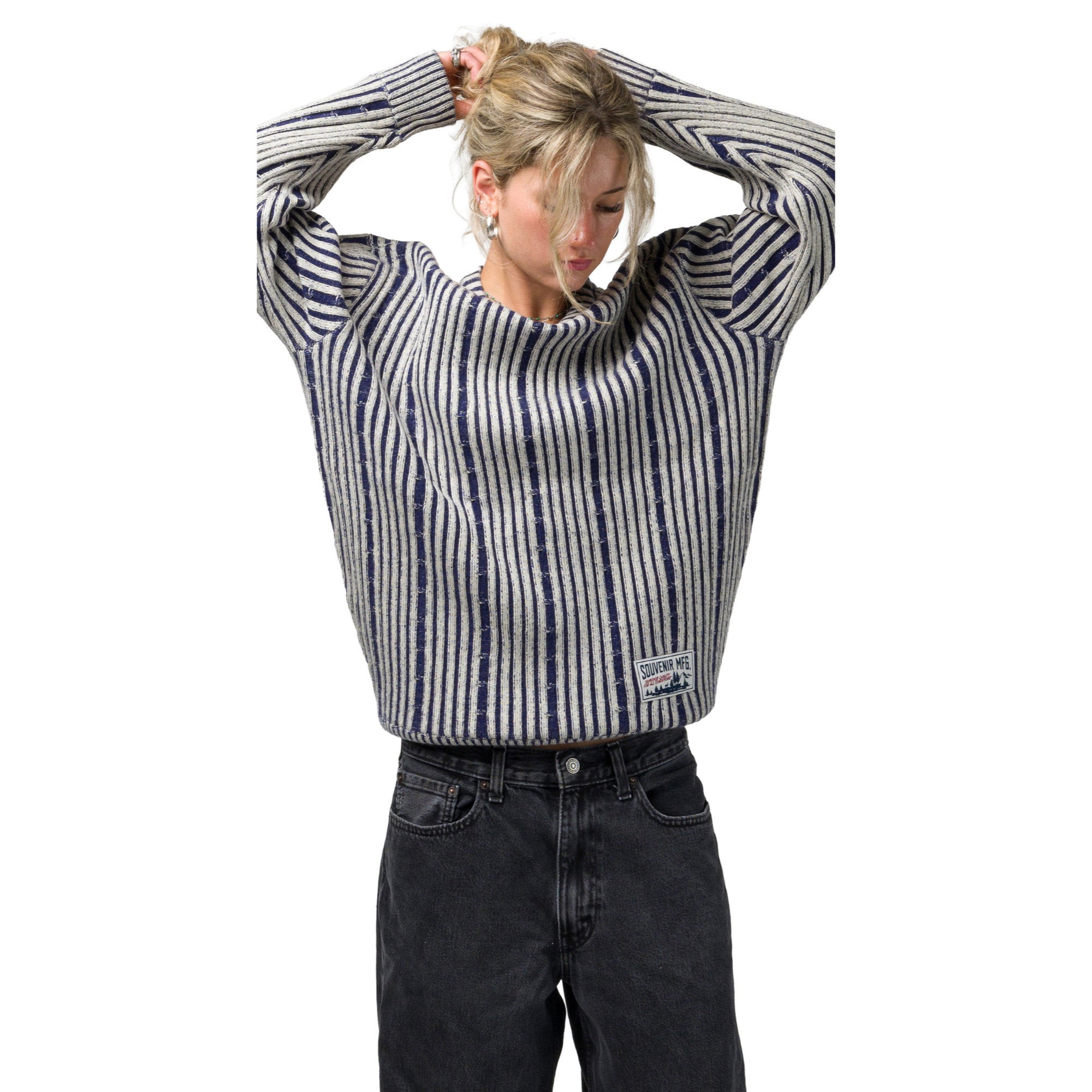 A blonder girl wearing a cropped, long-sleeve knit souvenir chunky sweater with vertical navy and off white stripes, paired with dark gray jeans, against a solid white background.